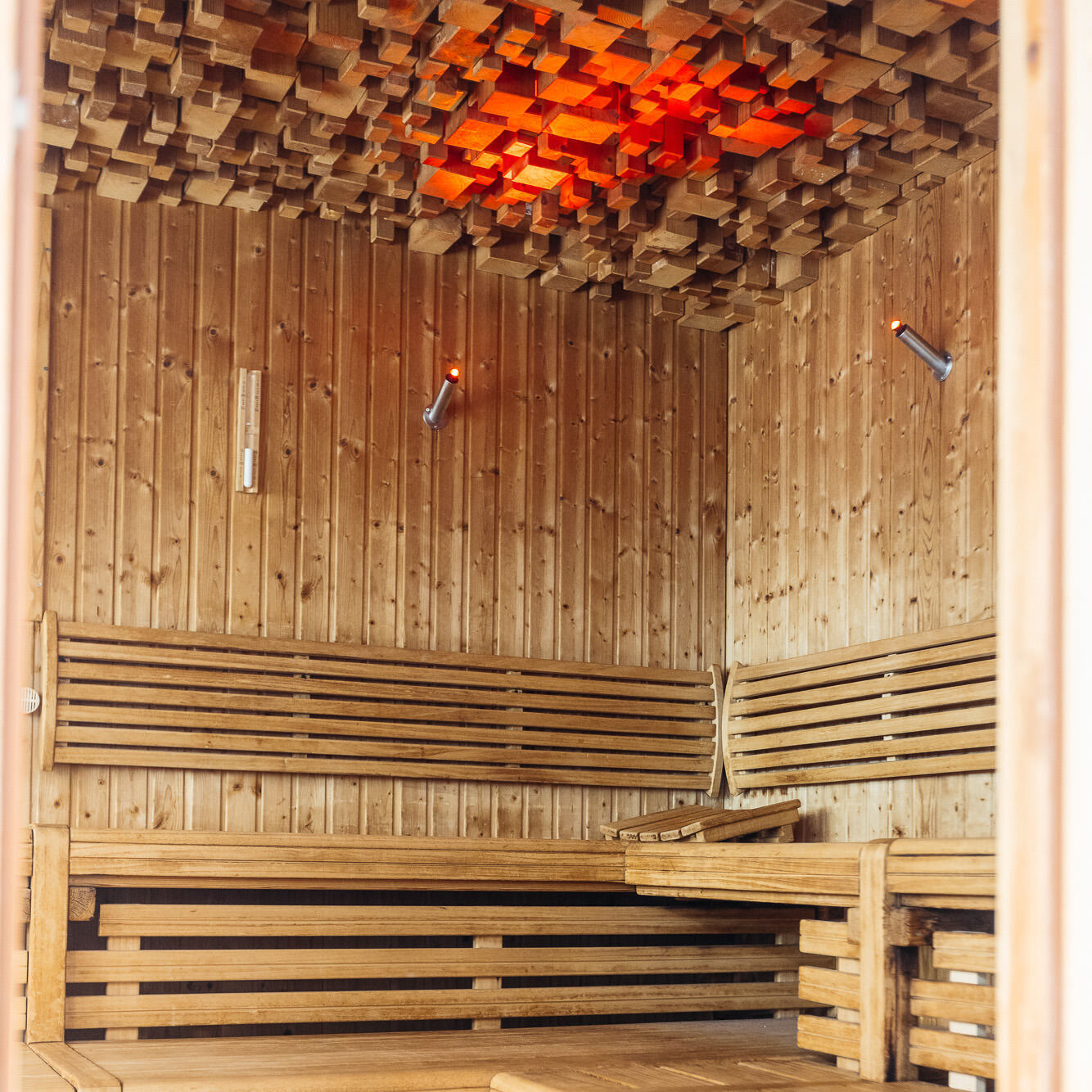 Eine Lampe aus vielen, kleinen Holzstempeln wirft ein helles rotes Licht in eine hölzerne, lichtduchflutete Sauna mit 3 Etagen .